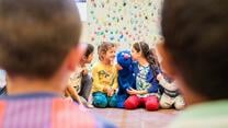 Refugee children sit on the ground in a circle playing witha puppet of the Sesame Street character Grover, a blue monster.